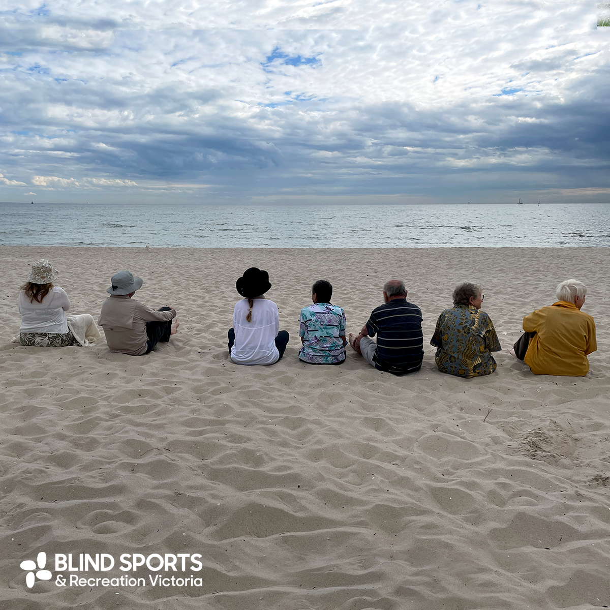 A slightly stormy looking sky above 7 people sitting on the beach facing away from the camera and looking towards the sea.